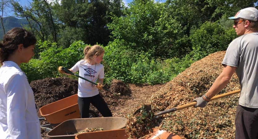 Two people shovel mulch into a wheelbarrow, held by another. 